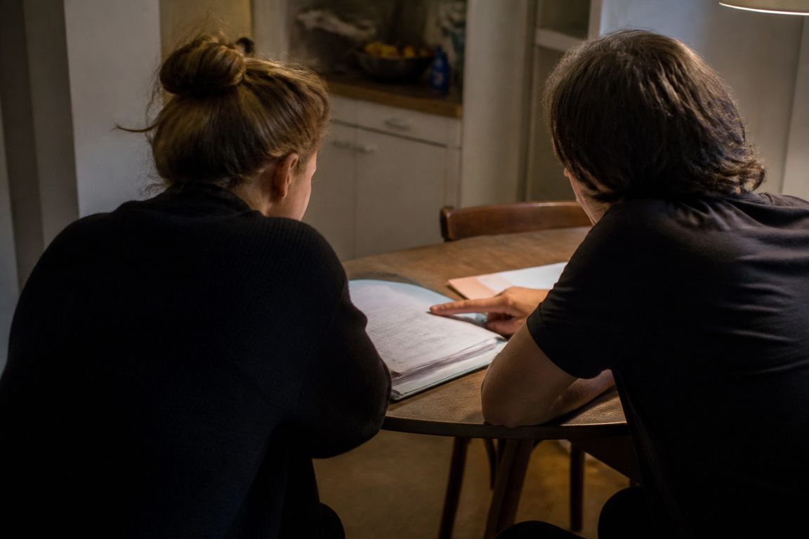 Zwei Personen sitzen von hinten gesehen an einem Küchentisch über einer Mappe mit Unterlagen, eine Hand zeigt auf eine Zeile