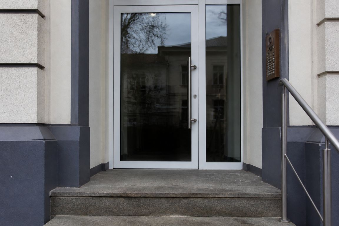 Die Glastür einer Arztpraxis von außen, daneben ein Klingeltableau aus Metall, davor eine abgetretene Steinstufe