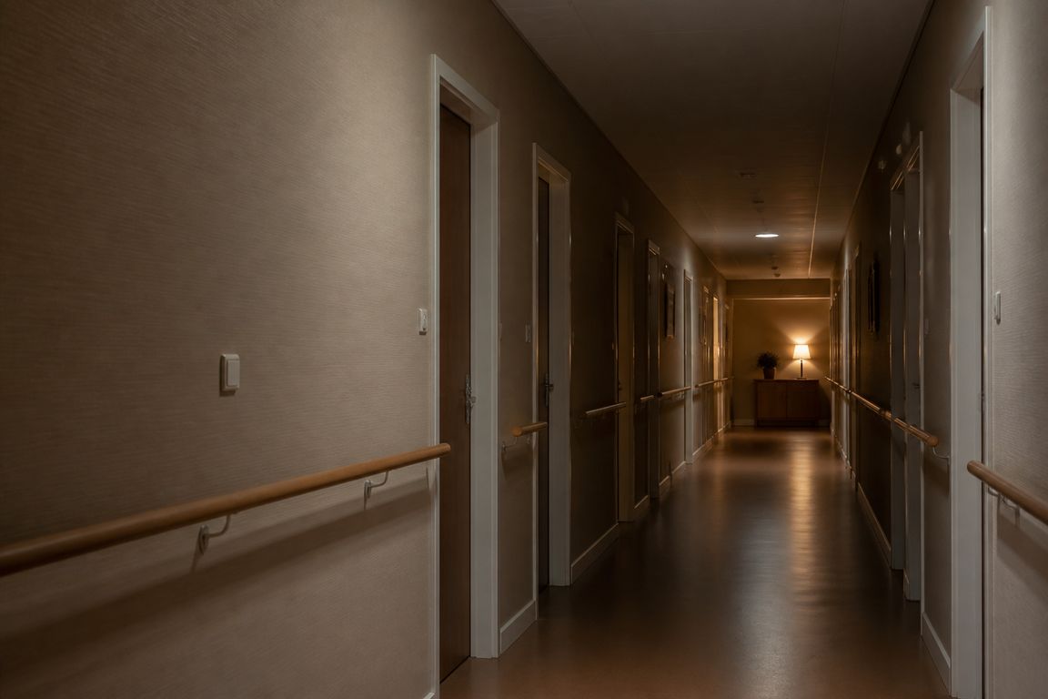 Ein langer stiller Flur in einer Pflegeeinrichtung mit einem Handlauf aus Holz, geschlossenen Zimmertüren und einer einzelnen Lampe am Ende