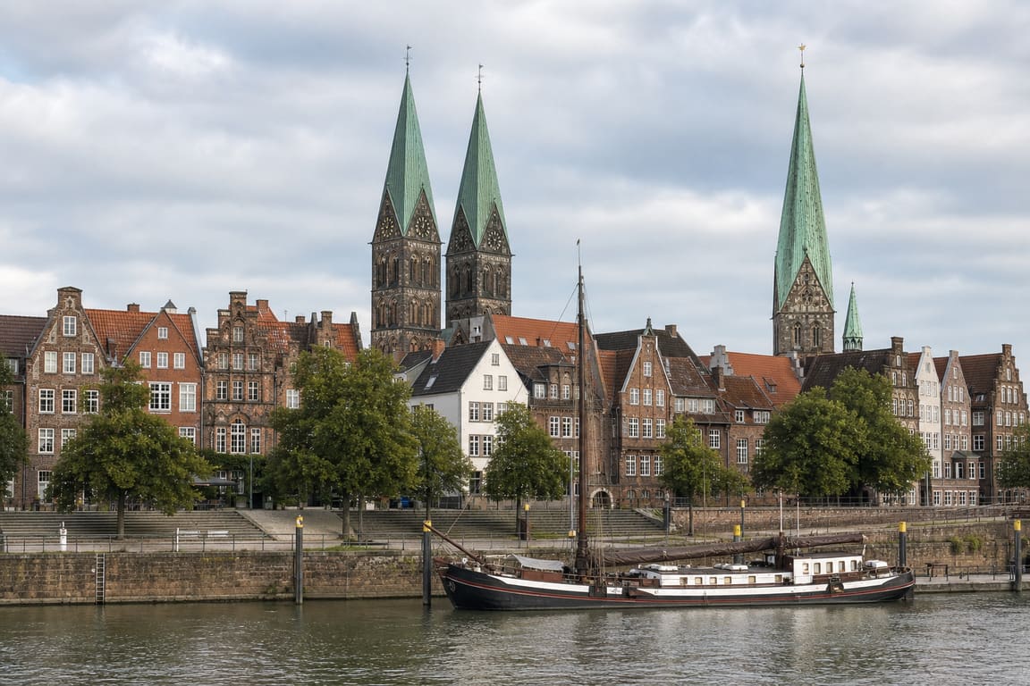 Hanseatische Altstadtfassaden mit Backsteingiebeln und zwei Kirchtürmen an einem Fluss, davor ein festgemachter Traditionssegler