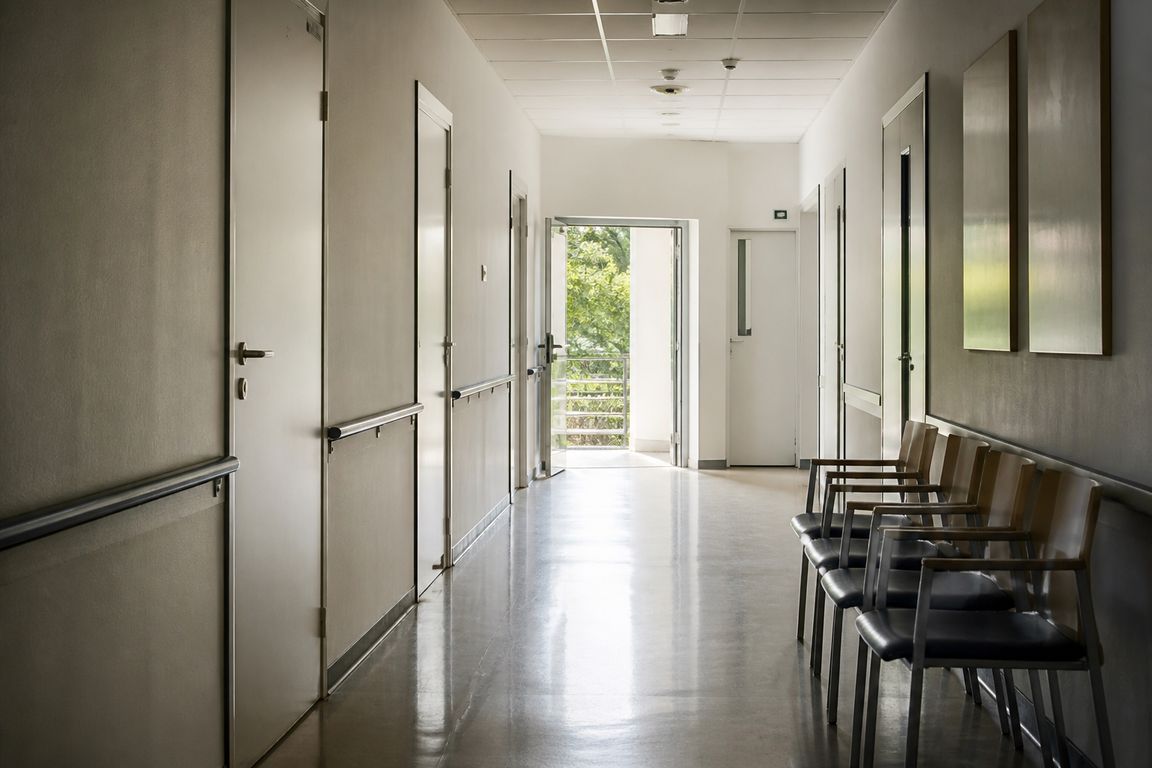 Ein leerer Krankenhausflur am Ende der Besuchszeit, an der Wand eine Reihe unbesetzter Wartestühle, am Ende eine offene Tür ins Tageslicht