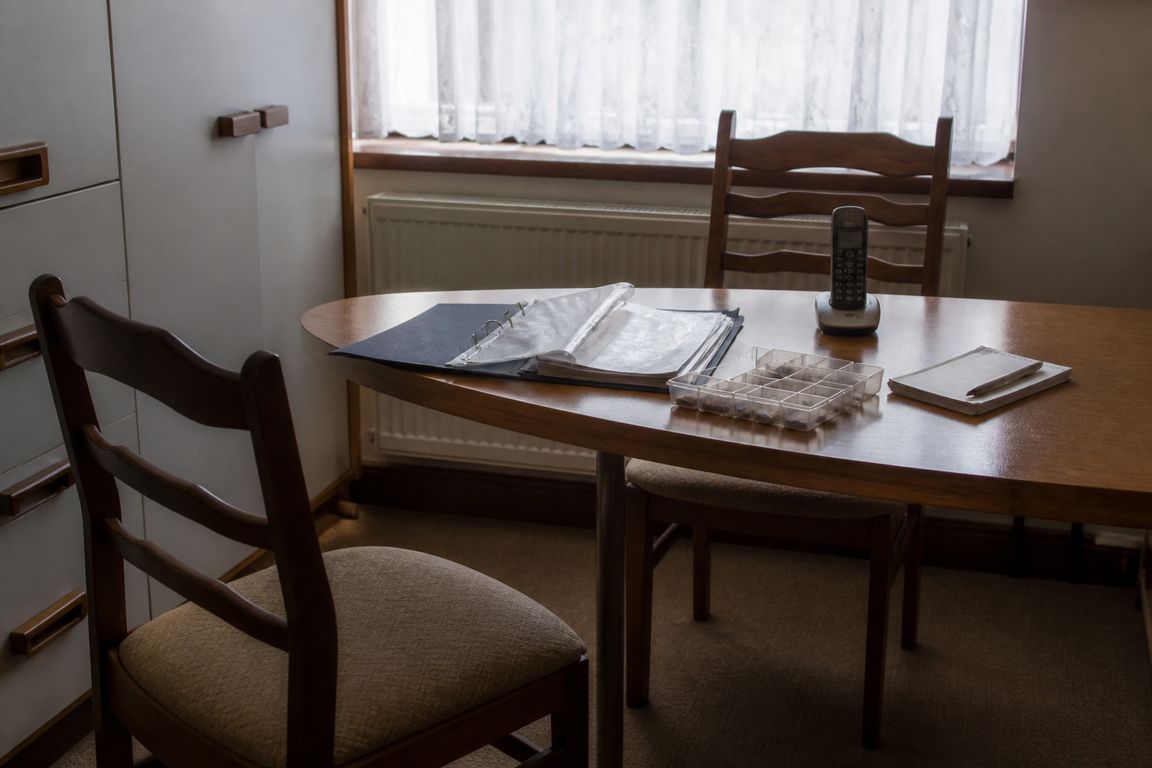 Ein Küchentisch in einer Wohnung mit einem aufgeschlagenen Ordner, einer Tablettenbox, einem Telefon und einem Notizblock, ein Stuhl ist herausgezogen